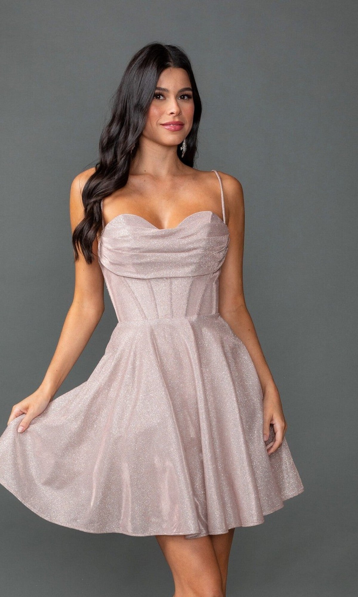 A woman stands against a gray background wearing the Dancing Queen 3421 short prom dress—light pink, sleeveless, with a glitter finish, fitted corset bodice, and flared skirt.